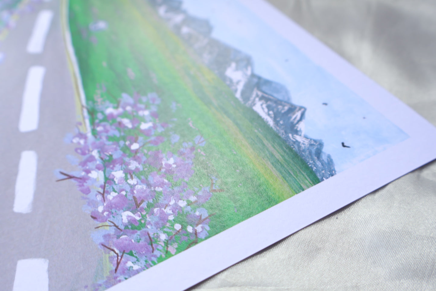 A roadside view, with a go ahead sign, and trees and flowers resembling bright purple lavender, giving off travelling vibes, with bright mountains further ahead upon a field of grass with bright pink and white flowers. Adding on to a clear blue sky, filled with birds viewed from a far in the background.