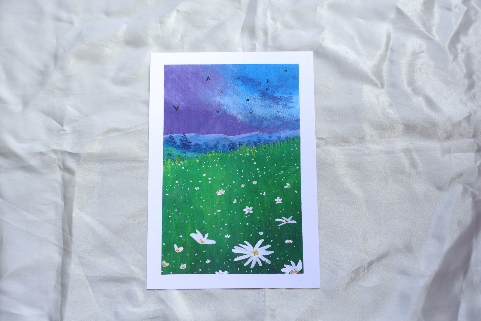 Purple bluey clouds under the dark evening blue sky, with soft mountains underneath it, covered with trees, followed by a bright foreground flowers, resembling daisies, glowing in the night sky, surrounded with yellowish and greenish tall grasses, meadow-like, and energetic birds flying in the sky.