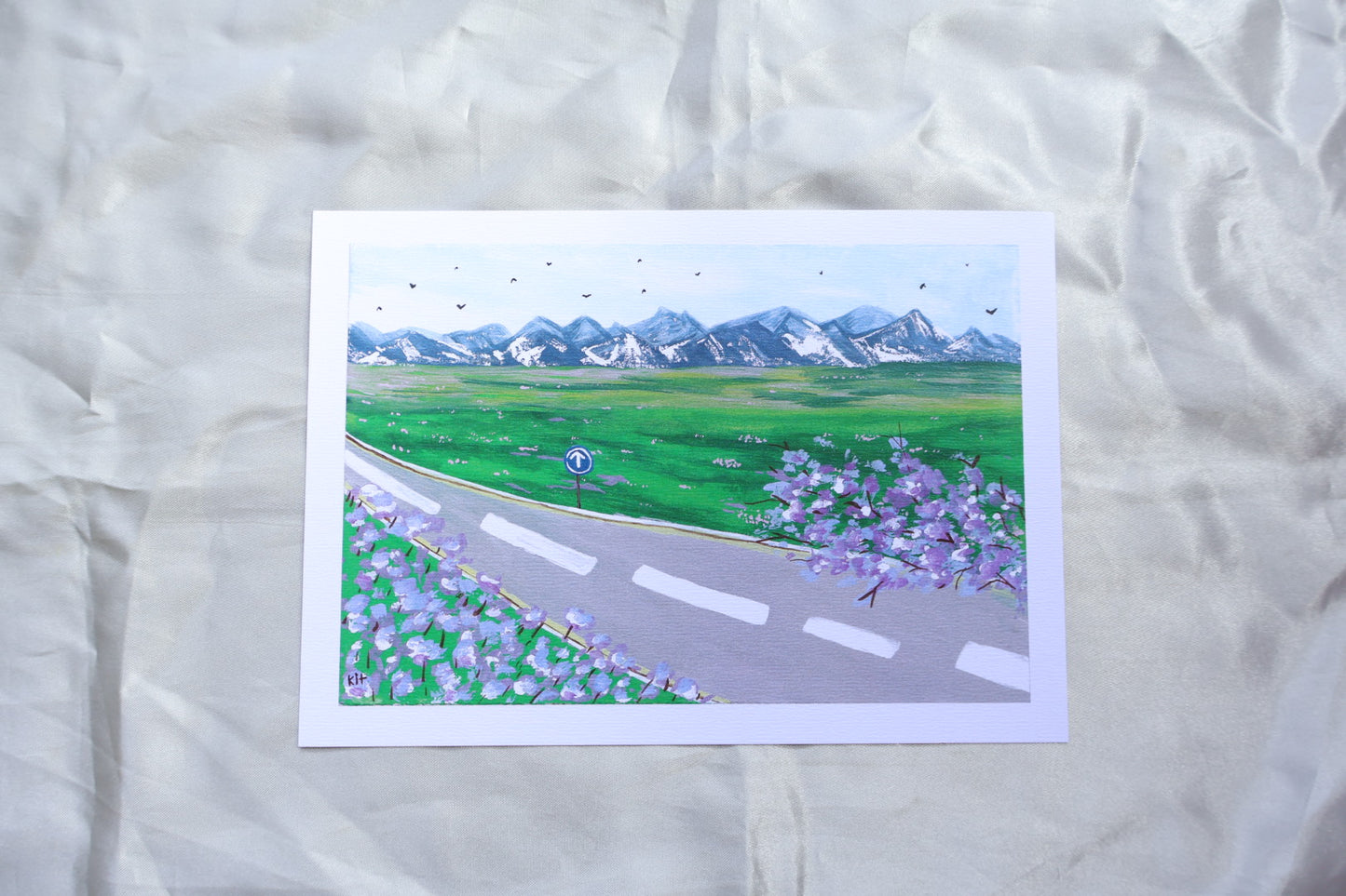 A roadside view, with a go ahead sign, and trees and flowers resembling bright purple lavender, giving off travelling vibes, with bright mountains further ahead upon a field of grass with bright pink and white flowers. Adding on to a clear blue sky, filled with birds viewed from a far in the background.