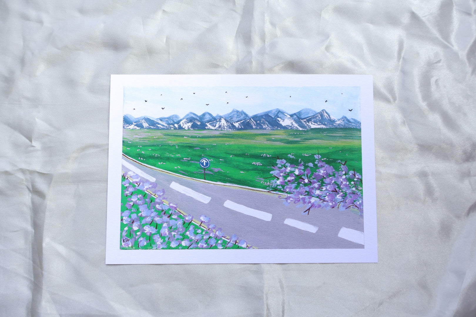 A roadside view, with a go ahead sign, and trees and flowers resembling bright purple lavender, giving off travelling vibes, with bright mountains further ahead upon a field of grass with bright pink and white flowers. Adding on to a clear blue sky, filled with birds viewed from a far in the background.