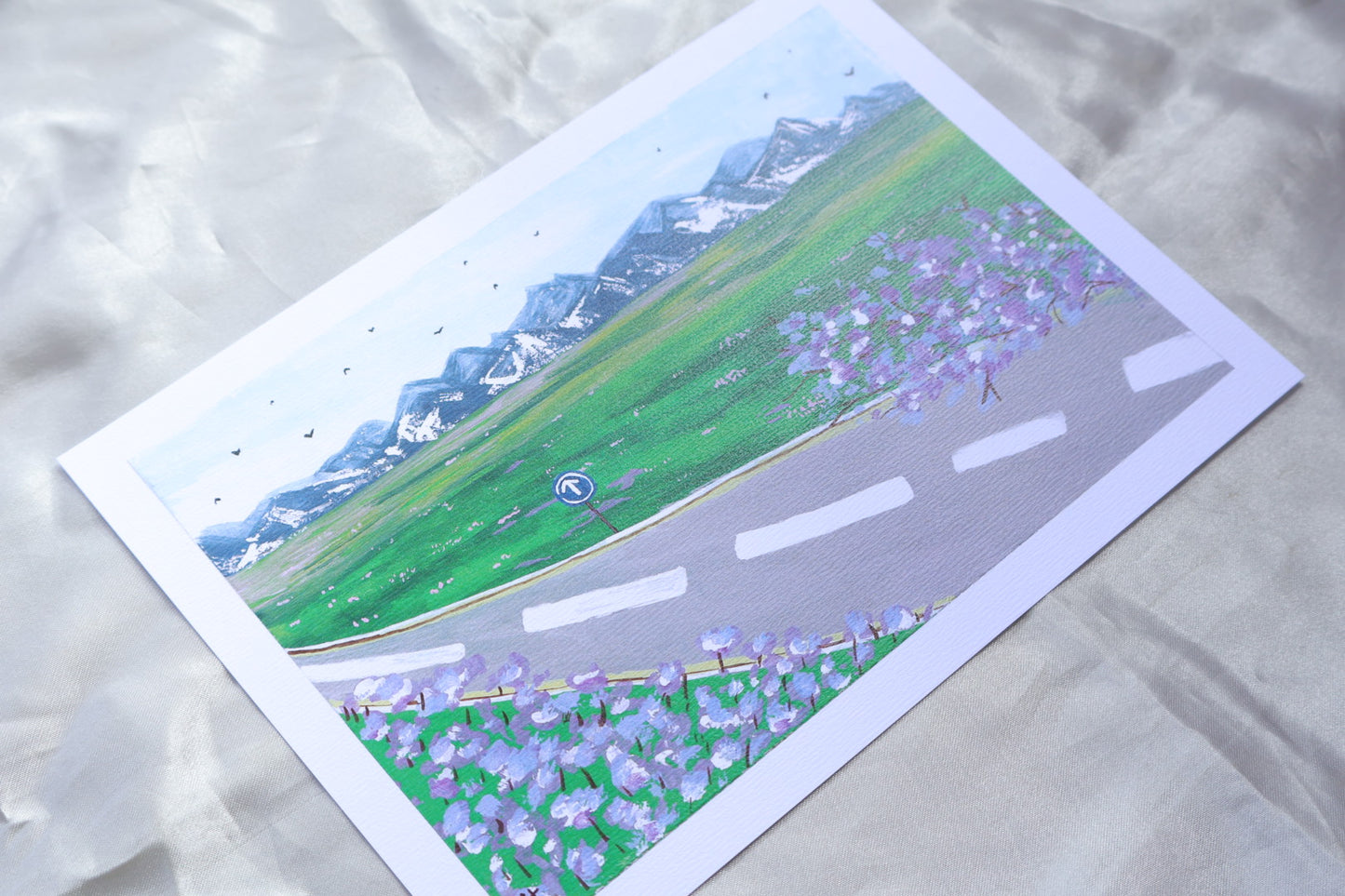 A roadside view, with a go ahead sign, and trees and flowers resembling bright purple lavender, giving off travelling vibes, with bright mountains further ahead upon a field of grass with bright pink and white flowers. Adding on to a clear blue sky, filled with birds viewed from a far in the background.